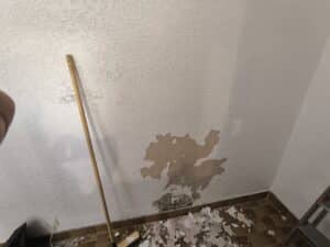 Mur blanc texturé écaillé révélant le plâtre, avec un balai et des débris de peinture au sol. Préparation rénovation.