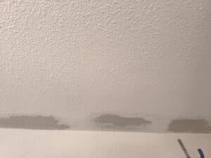 Plafond texturé en relief style popcorn avec des zones grises où la texture a été retirée ou réparée, près du mur. Une perche est visible.