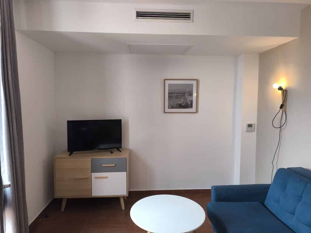 Salon moderne avec canapé bleu, table basse ronde, meuble TV bois clair/gris/blanc et TV. Mur blanc, sol carrelé, climatisation visible.