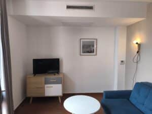 Salon moderne: canapé bleu, table basse blanche, meuble TV Schneider, cadre art marin N&B, lampe murale, thermostat Panasonic, rideaux gris.