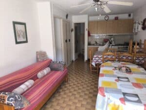 Studio cosy: canapé-lit rayé, coin repas avec tables colorées, cuisine bois équipée. Sol carrelé. Un magazine Gala est sur la table.