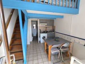 Studio moderne avec escalier en bois, mezzanine lit, cuisine équipée grise et table à manger avec chaises. Mur carrelé bleu.