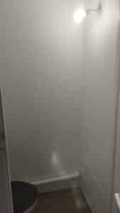 Petite pièce aux murs texturés blancs, éclairée par une ampoule nue suspendue. Un siège de toilettes sombre est visible en bas à gauche.