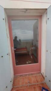 Réflexion d'une personne se photographiant dans une porte vitrée rose ouverte sur une chambre simple avec lits et vue sur mer et plage, sous un ciel nuageux.